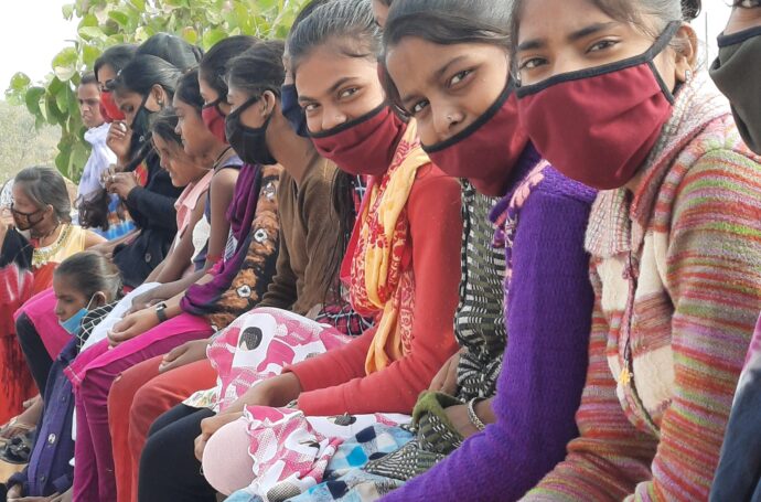 Long line of Indian girls in facemasks, sitting, looking towards camera on their left.