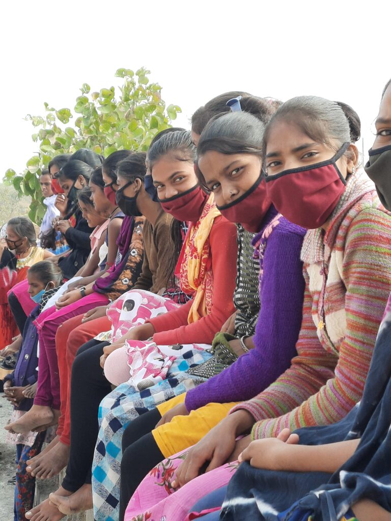 Long line of Indian girls in facemasks, sitting, looking towards camera on their left.