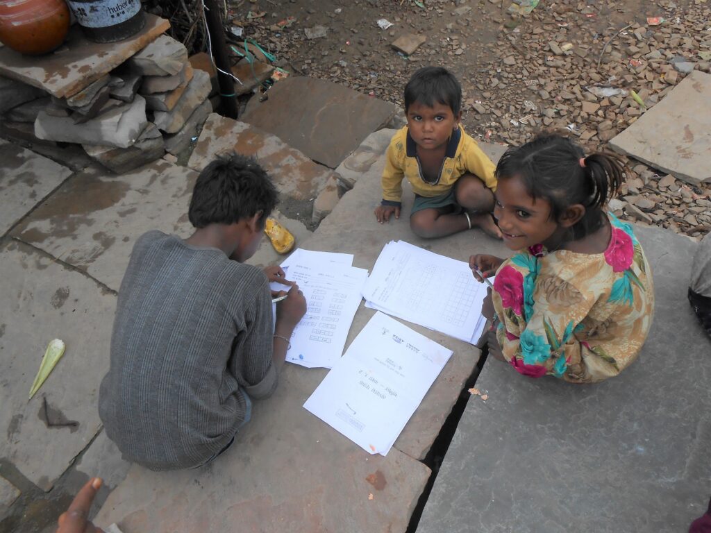 Group of 3 children of different ages, sitting on stone slabs, with worksheets spread around. in Budhpura, Rajasthan, India. They're not in school because of Covid restrictions.