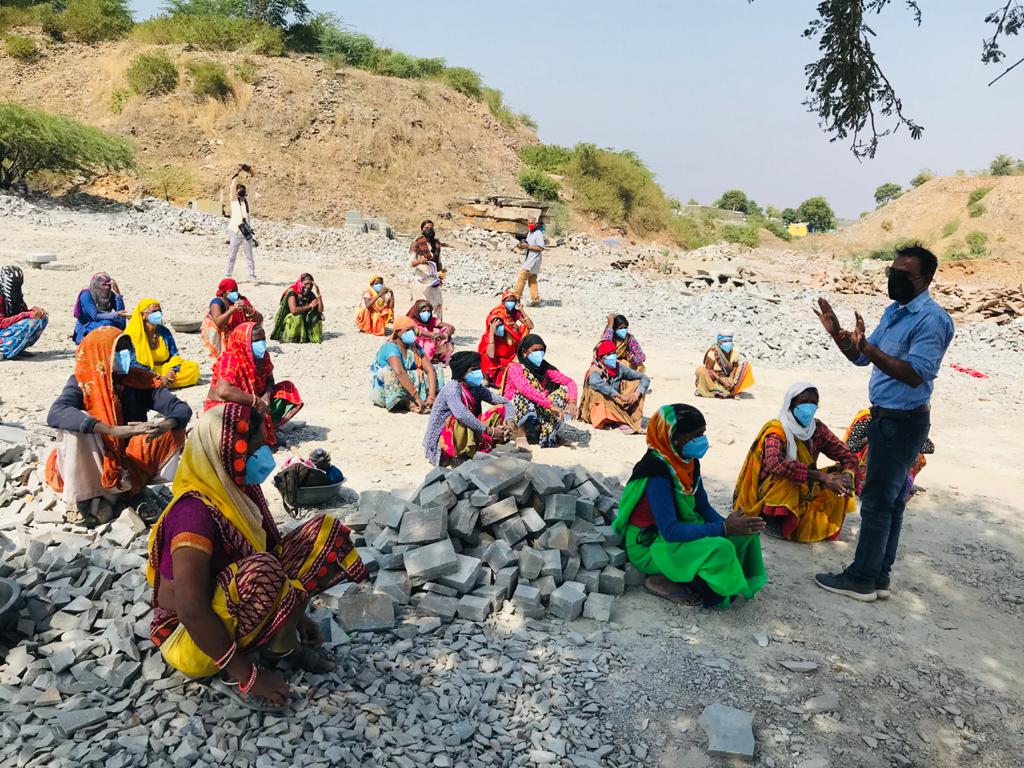 Women sitting spaced apart in rows on dusty ground receiving instructions. 